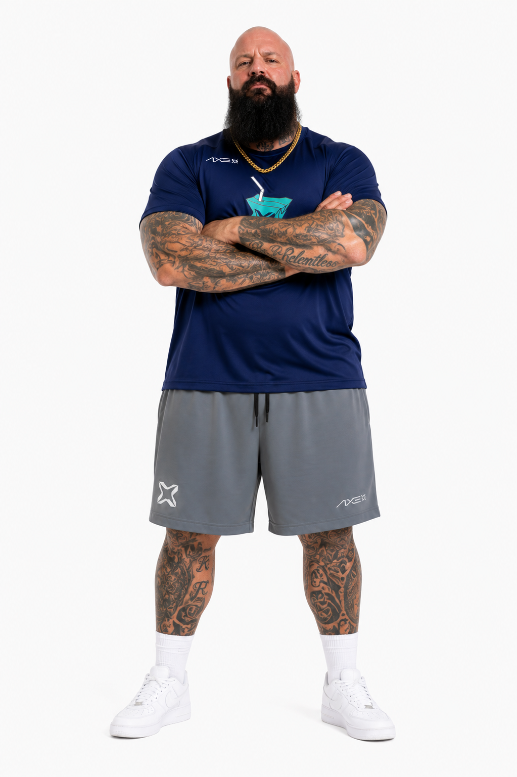 A man with a full beard and tattoos stands with arms crossed, wearing a navy blue athletic t-shirt with a teal logo, gray athletic shorts, white socks, and white sneakers, against a plain white background.