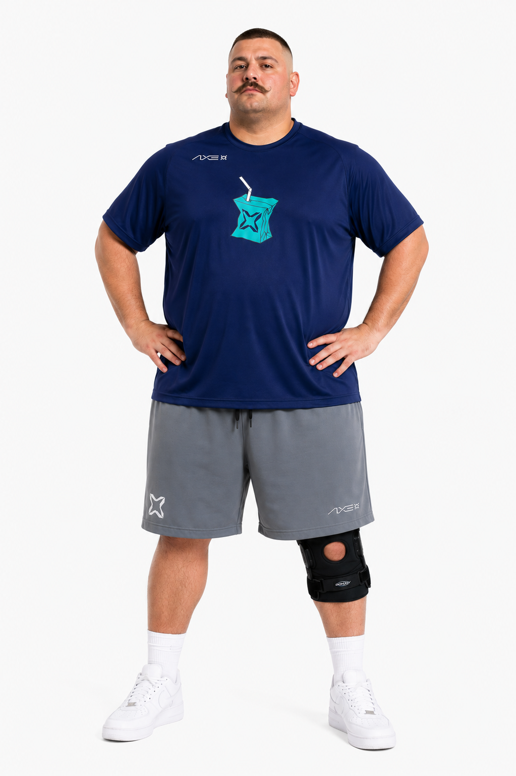 A man modeling a navy blue athletic t-shirt with a teal juice box graphic, gray athletic shorts, a black knee brace, and white sneakers against a light gray background.