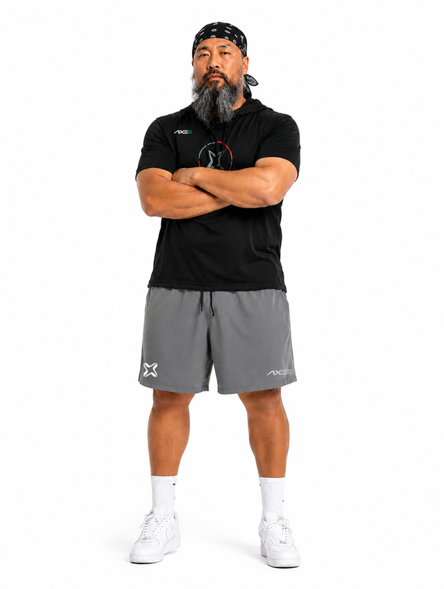 A man with a beard and bandana stands with arms crossed, wearing a black short-sleeve athletic shirt, gray athletic shorts, white socks, and white sneakers against a plain white background.