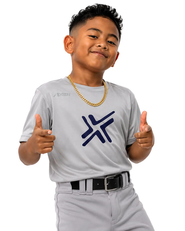 A young boy wearing a light gray baseball uniform with a navy blue stylized 'X' logo on the chest, a gold chain necklace, and a black belt, posing with thumbs up against a white background.