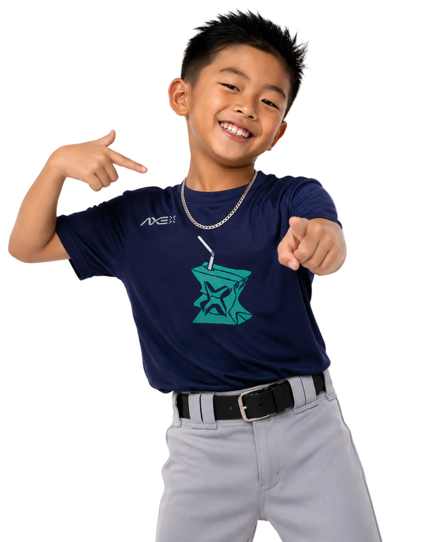 A young boy smiling and pointing at a navy blue t-shirt with a teal graphic on the front, paired with gray pants and a silver chain necklace, against a plain white background.