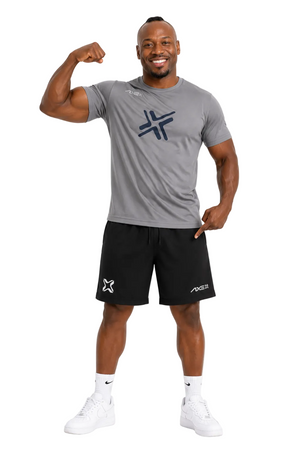 A man modeling a gray short-sleeve athletic t-shirt and black athletic shorts with white sneakers, posing with one arm flexed against a light gray background.