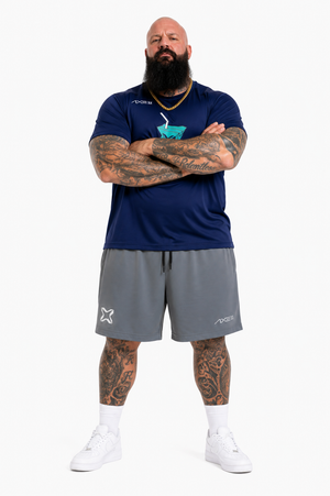A man with a full beard and tattoos stands with arms crossed, wearing a navy blue athletic t-shirt with a teal logo, gray athletic shorts, white socks, and white sneakers, against a plain white background.